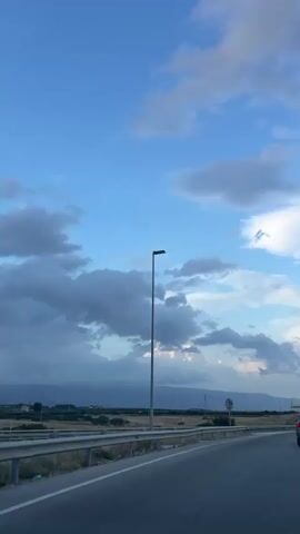 This is a vertical, slightly shaky, first-person perspective video of a car driving on a highway. The sky is blue with scattered white and gray clouds. A tall lamppost is visible on the left side of the road. In the distance, there are mountains and fields. Several other cars are visible on the highway, including a silver SUV in front of the viewpoint vehicle. The sun appears to be setting or is low in the sky, casting a soft light.