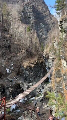The video captures a scenic view of a deep gorge in Arshan, Russia, featuring a series of suspension bridges spanning a flowing river. Several people are seen walking across the wooden plank bridges and navigating the rocky terrain.

In the foreground, a wooden structure with stairs and railings serves as an access point to the main suspension bridge, where a few individuals are standing. The primary suspension bridge, constructed from wooden planks and supported by cables, extends across the gorge. Further along, another section of a similar bridge can be seen.

The gorge walls are steep and rocky, covered with trees that are mostly bare, indicating either late winter or early spring. Patches of snow are visible on the ground and rocks, contrasting with the green-tinged water of the river below. The sky is clear and blue, suggesting a sunny day. The overall scene depicts an active outdoor environment, likely a popular hiking or tourist destination, where visitors are exploring the natural landscape and the unique bridge structures. No visible text is present in the video.