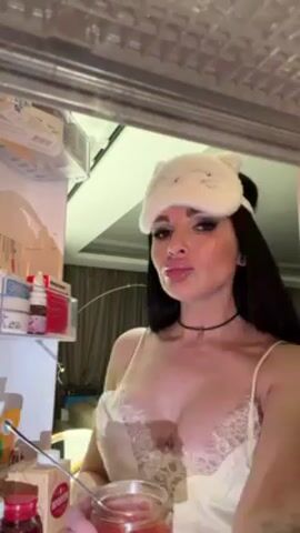 The video shows a woman standing in front of an open refrigerator. She wears a white sleep mask with cat ears, a light-colored lace-trimmed slip, and a choker necklace. She has long dark hair, heavy makeup, and large lips. She holds a small jar of red caviar in her left hand and a spoon in her right. She eats a spoonful of caviar, looking directly at the camera, then licks her lips. The refrigerator contains various food items, including cartons, bottles, and small boxes of what appear to be medications. The environment is dimly lit with a small string of lights visible in the background.