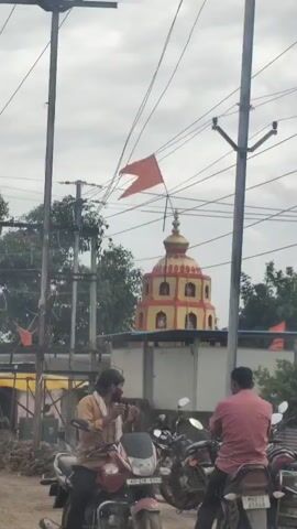 The video shows a religious structure with a tiered, domed roof and red and yellow decorative elements. An orange flag is visible flying from a pole in front of the structure, and another smaller orange flag is on a lower pole to the left. Several utility poles with numerous wires crisscross the frame. In the foreground, a person sits on a motorcycle, their face partially obscured, and they appear to be adjusting something near their face. Another person in a light-colored garment stands with their back to the camera on the left side of the frame. Debris and construction materials are scattered on the ground. The sky is overcast.