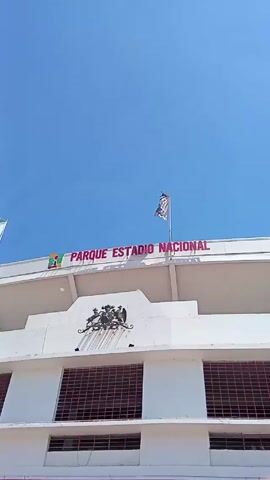 The video shows a partial view of the exterior of the Estadio Nacional Julio Martínez Prádanos, a national stadium located in Santiago, Chile. The building is predominantly white, with the words "PARQUE ESTADIO NACIONAL" written in red lettering across the top. There are flags flying atop the building. Below the sign is an emblem and barred windows. The words "BIENVENIDOS" are on a pink banner. A man wearing a white t-shirt and a baseball cap walks on a track and points his left arm. The sky is blue.