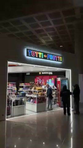The video shows the exterior of a Tutti-Frutti shop in what appears to be an airport. The sign "TUTTI-FRUTTI" is in large, brightly colored letters. Below that is the text "BARCELONA DUTY FREE". Inside the shop, various shelves and displays hold confectionery and other items. A man with gray hair is browsing the products. A person in a dark shirt is standing near a refrigerator. The environment looks like an airport terminal with a modern ceiling design.