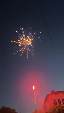 A low-angle vertical shot shows fireworks exploding in the night sky. The fireworks, in yellow and orange hues, burst into various patterns, illuminating parts of the dark sky. A bright red streak of light, indicative of a firework launching, ascends from the bottom right. In the bottom right corner, a building with red-tiled roofs and windows is partially visible, providing a contrasting backdrop to the aerial display. In the bottom left, the tops of trees can be seen. Two thin, dark lines, possibly power lines, stretch diagonally across the frame, dividing the sky. Faint stars are scattered across the dark sky.