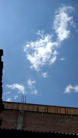 A low-angle shot of a clear blue sky with scattered white clouds. To the right, a brick wall with wooden beams and rebar is visible. In the foreground, a dark silhouette of a roofline or overhang is present. The lighting suggests daytime.