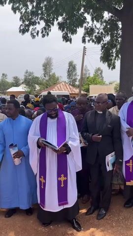 Sure, here's a description of the video:

The video captures a gathering of individuals outdoors, likely attending a ceremony or service. A central figure, dressed in a white clerical robe with a purple stole adorned with gold crosses, is seen reading from a book. He is wearing sunglasses.  He is surrounded by other clergy members in similar attire, as well as people dressed in more casual wear. They stand closely together. A tree with ample foliage provides some shade, and in the background, there are houses, vehicles, and additional attendees. The general atmosphere suggests an outdoor religious event or memorial.