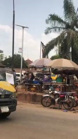 The video shows a street scene with several commercial vehicles, including a yellow van and a three-wheeled auto-rickshaw. The foreground features the side view of the yellow van with its front heavily damaged. The vehicle is moving, passing by a busy roadside market area. Several market stalls are set up under umbrellas, with vendors and potential customers visible. One individual is sitting on the edge of a table within one of the stalls. A motorcycle is parked near the market. The backdrop includes a palm tree and a pole with a banner attached. The general setting appears to be a sunny day.