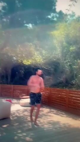 This media file shows a man in dark patterned swim trunks standing on a wooden deck outdoors. He is shirtless and has long dark hair pulled back, with a beard. He is gesturing with his hands and appears to be talking or singing. In the background, there is a wooden fence, lush green trees and foliage, and two large, light-colored beanbag chairs on the deck. The sunlight suggests it is daytime, likely with clear weather. A hand holding up two fingers in a peace sign is visible in the foreground on the right side of the frame. There is no visible text in the media file.