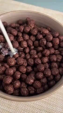 A close-up shot shows milk being poured from a container into a bowl of chocolate cereal. The cereal pieces are small and round, and the milk is creating a creamy white swirl as it mixes with the chocolate. The bowl is light brown and sits on a textured mat. The background is out of focus, but appears to be a table with a light blue surface.