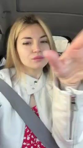 A blonde woman wearing a white jacket and red floral dress is sitting in a car with her seatbelt on. She is looking at the camera and waving. There is music playing in the background. The car appears to be in motion.