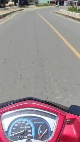 The video shows a first-person perspective of someone riding a red scooter on an asphalt road. The scooter's dashboard is visible at the bottom of the frame, displaying a speedometer, fuel gauge, and odometer showing 17533 km. The road has a yellow double solid line in the middle and a white broken line on the right shoulder. On the left side of the road, there are several buildings, some with corrugated metal roofs. Utility poles with power lines run along both sides of the road. Another scooter, red in color, is seen approaching from the opposite direction and then passing on the left. The sky is clear with a few white clouds, and the weather appears sunny. The road curves slightly to the right in the distance.