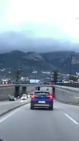 The video depicts a road scene from a vehicle's perspective. The camera is behind a blue Renault car, driving on a road with multiple lanes. Overhead, there is a concrete bridge with several archways, beneath which the road continues. Traffic flows both on the road the camera vehicle is on and beneath the bridge. The background features a view of mountains shrouded in grey clouds, with palm trees lining the roadside. Streetlights are visible along the bridge. The overall atmosphere appears overcast and muted.