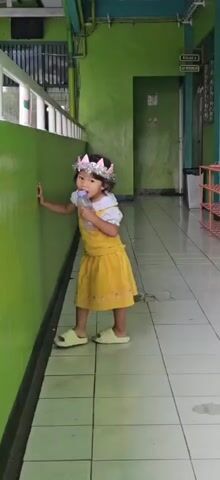 A young girl with a crown on her head stands in a hallway and drinks from a bottle. The hallway has green walls and tiled floors. A trash can and some shelves are visible in the background.