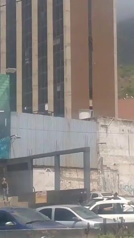 This video appears to be taken from a moving vehicle on a street in Maiquetía, Venezuela. The scene is urban, with a mix of buildings, some appearing modern and others more dilapidated, situated on a hillside.

In the foreground, a blue car is visible, followed by a white car. The video pans to show a street with parked vehicles, including a pickup truck. A person can be seen walking on the sidewalk in the distance. Further down the street, a white van is parked.

The background features a mountainous landscape, with many houses built on the slopes, suggesting a densely populated area. The architecture varies, with some multi-story buildings and what appear to be more informal constructions. The weather seems to be clear and sunny, indicating it is daytime.

A woman in a white t-shirt and patterned pants is visible on a raised area, looking towards the left. The overall impression is a snapshot of everyday life in a somewhat hilly, urban environment in Venezuela. There is some visible graffiti on a wall in the background, but no specific text is legible.