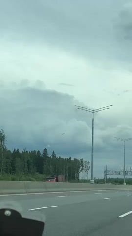 The video shows a view from a moving vehicle on a highway. The sky is overcast with dramatic clouds. On the left side of the highway, there is a dense forest. A red truck is visible on the left shoulder of the road. Tall streetlights with multiple lamps line the highway. An airplane is visible in the sky between two streetlights. On the right side of the highway, there is a large overhead sign structure, and several vehicles, including a white camper van and a dark car, are driving ahead. The camera is positioned from the perspective of a passenger in the vehicle, with a portion of the vehicle's interior visible in the foreground at the bottom left of the frame.