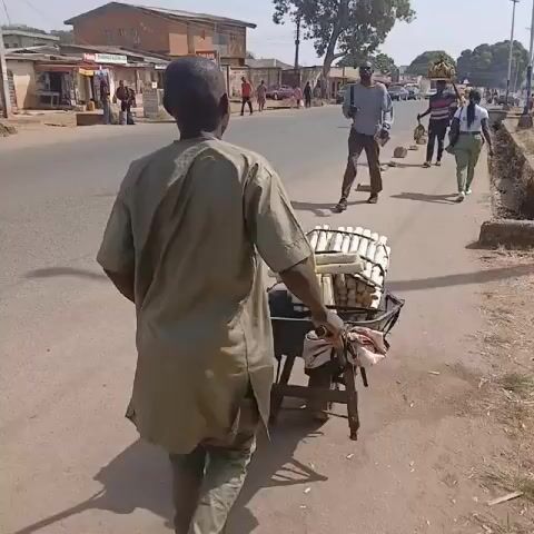The video shows a street scene in what appears to be a suburban area.

A man in a long, light-colored shirt is pushing a small cart loaded with eggs along the side of the road. Several other people are walking on the same side of the street, including a man in a grey shirt and another man carrying a tray of plantains on his head.

Across the street, there are several small businesses and people standing outside. The overall environment seems sunny and relatively calm.