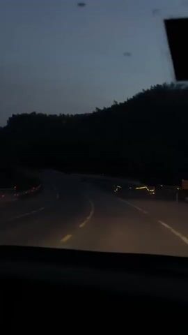 The video shows a view from inside a vehicle, looking out through the windshield at a dark road winding through a forest. The sky is dark blue, indicating it is either dawn or dusk. The headlights of the vehicle illuminate the road ahead, revealing yellow lane dividers and guardrails on either side of the road. The trees on the hills are silhouetted against the sky, appearing very dark. There are no other vehicles visible on the road. The camera is stationary, suggesting the video is a recording of a continuous drive.