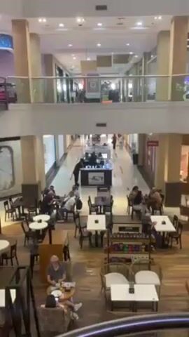 From a high vantage point, the video captures an indoor scene of a mall's interior. The ground level features a coffee shop seating area with numerous tables and chairs occupied by people. Most are seated and appear to be engaging in conversation or using their phones.  Above, a second level balcony overlooks the area. Along the balcony's edge are shops or storefronts. In the mall's corridor on the second level, a small retail kiosk is visible, with people gathered around it. The mall has a clean, modern design with neutral tones and ample lighting.