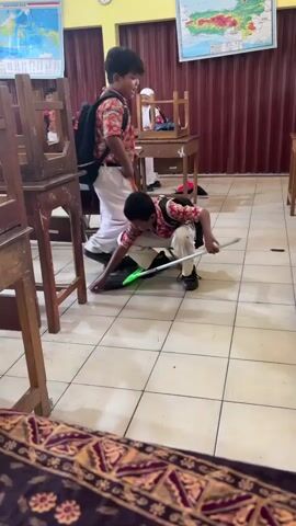 Two young boys are in a classroom, sweeping the tiled floor. One boy, wearing a red and white patterned shirt, white pants, and a backpack, is in a crouched position, sweeping with a green-bristled broom and a dustpan. The other boy, also wearing a similar patterned shirt and pants, and a backpack, is standing and sweeping with a broom. The classroom contains wooden desks and chairs, some of which are stacked. Maps and educational charts are displayed on the walls. The overall environment appears to be a school setting during the day.