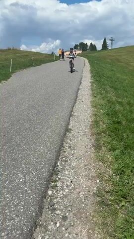A person wearing a helmet and a tank top is riding a bicycle on a paved path. The path is surrounded by green grass and there are trees in the background. The sky is blue with some clouds. There are other people in the background walking on the path. The person on the bicycle is moving towards the camera.