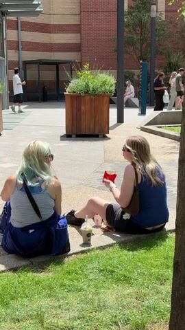 Two women sit on the ground, facing each other, in an outdoor paved area with grass. The woman on the left has blonde hair with streaks of light blue and wears a striped tank top, a blue jacket, and blue pants. She has a blue bag on her right side and headphones on. She holds a phone in her right hand. The woman on the right has blonde hair and wears a blue tank top and black shorts. She holds a red carton of french fries and is eating from it. A cup with a straw is on the ground between them. In the background, a person in a white shirt and black shorts walks away from the camera, and another person sits on a bench. A planter with green plants is in the middle ground. A brick building is behind the planter, and a red post box is visible on the right. The scene is illuminated by daylight, indicating it is daytime.
