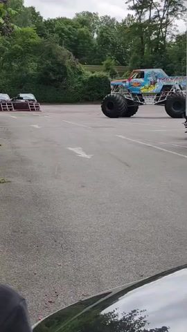 A monster truck is shown in a parking lot. The truck is blue and yellow and has very large tires. There are two cars parked on the left side of the parking lot and another monster truck in the background. The ground is asphalt and there are trees and bushes in the background. The sky is cloudy. The video is shot from a low angle, looking up at the monster truck.