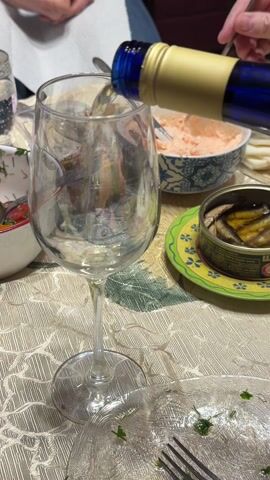 A person pours white wine from a blue and gold bottle into a wine glass. The table is set with bowls of food, including a pink dip, a small salad, a can of smoked fish, and pita bread. A plate with food remnants and a fork is also visible. The tablecloth has a light-colored, textured pattern. Another person is partially visible in the background, with their hands clasped.