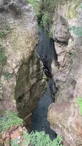 The video shows a narrow gorge with steep, rocky walls. A stream of water flows through the bottom of the gorge. The water is dark blue and appears to be moving quickly. There is some vegetation growing on the sides of the gorge, including trees and bushes. The light is dim, suggesting that the gorge is deep and shaded. There is a small wooden structure visible on the left side of the gorge, possibly a platform or a bridge. The perspective is from above, looking down into the gorge.