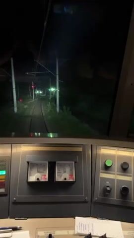 The video captures the interior of a train cab at night. The camera pans from the front view of the tracks ahead, which are illuminated by the train's headlights, to the right side of the cab, showing various control panels with gauges and screens. The exterior environment is dark, with glimpses of trees and overhead power lines. A yellow sign with the letter "R" and a star symbol is visible on the interior ceiling.