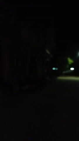 The video shows a dimly lit outdoor scene at night. A street light illuminates a paved pathway. On the right side of the pathway, there is a two-story building with orange and green accents. Some windows are visible, and a doorway is open, revealing an interior with light. Several parked motorcycles are visible in front of the building. On the left side of the pathway, there are some trees and the dark outlines of other structures. The overall impression is a quiet, possibly deserted, area at night, likely a school or public facility given the architecture and layout.