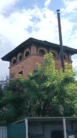 A short video clip shows an old brick structure with arches and a chimney on a bright, cloudy day. In the foreground, there are some green metal bins and a fence. The camera pans to the right, revealing more of the sky and a partially built house with a blue roof in the distance. Trees and foliage surround the brick structure and the partially built house.
