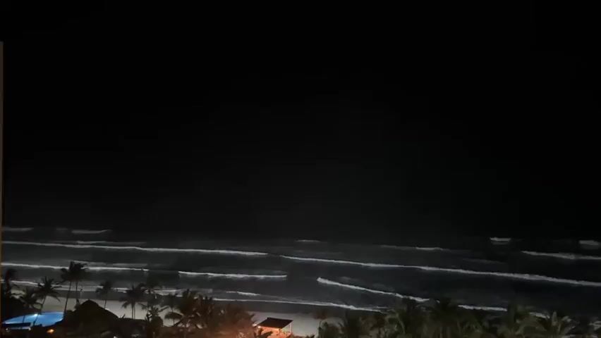 A nighttime drone view shows large white waves crashing on a dark beach. The beach is lined with palm trees. Bright lights are visible on the beach.
