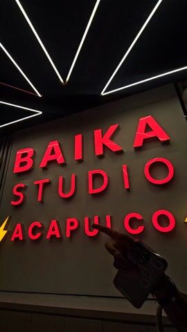 A close-up, low-angle shot shows a sign that reads "BAIKA STUDIO ACAPULCO" in large, illuminated red letters. The sign is mounted on a gray background. Above the sign, a geometric arrangement of white, thin neon lights hangs from the dark ceiling. A small, yellow lightning bolt graphic is visible to the lower left of the word "ACAPULCO." Part of a person's arm is briefly visible in the lower right corner of the frame. The lighting suggests an interior location. The camera is slightly tilted upwards.