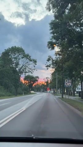 The video shows a view from inside a car driving on a wet road. The sky is cloudy and there is a sunset with pink and orange hues visible in the distance. There are trees lining the road on both sides, and streetlights are on. There are some traffic lights visible, and road markings are present on the asphalt. A few cars are visible in the distance. The overall impression is that of a scenic drive during sunset.