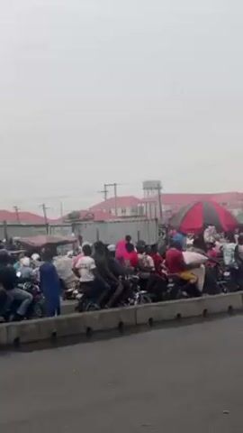 The video shows a busy street scene in an urban area. The camera is likely inside a moving vehicle, as the perspective shifts.  A large group of people, many on motorcycles, are gathered near a sidewalk, creating a crowded scene.  Several yellow buses are present, one prominently labeled "LASGHEY".  There are also cars on the road, including a gray sedan. Street vendors are visible with umbrellas.  The overall environment appears congested and bustling with activity. The weather appears overcast.