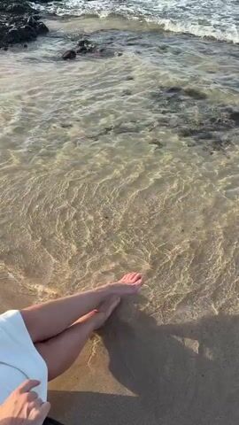 A person is seen from a high angle, sitting on the sandy shore at Al Fiqait, United Arab Emirates. Their bare feet are resting in the wet sand as small, clear waves gently wash over them and recede. The water is translucent, revealing the sandy bottom. In the background, calm ocean waves are visible, with dark rocks or coral formations peeking out of the water further from the shore. The light suggests it is daytime, possibly late afternoon, with soft shadows cast on the sand. The scene conveys a sense of tranquility and relaxation, with the person enjoying the calm beach environment. No text is visible in the video.