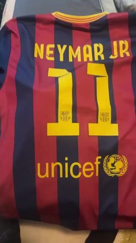 The video shows a close-up, static shot of the back of a Neymar Jr. FC Barcelona soccer jersey. The jersey has blue and red vertical stripes. In yellow lettering, the name "NEYMAR JR" appears at the top, followed by the number "11" printed twice with a small FC Barcelona logo inside each numeral. Below the numbers is the word "unicef" and a UNICEF logo. The jersey lies on a surface with folded clothing or a blanket visible in the background.