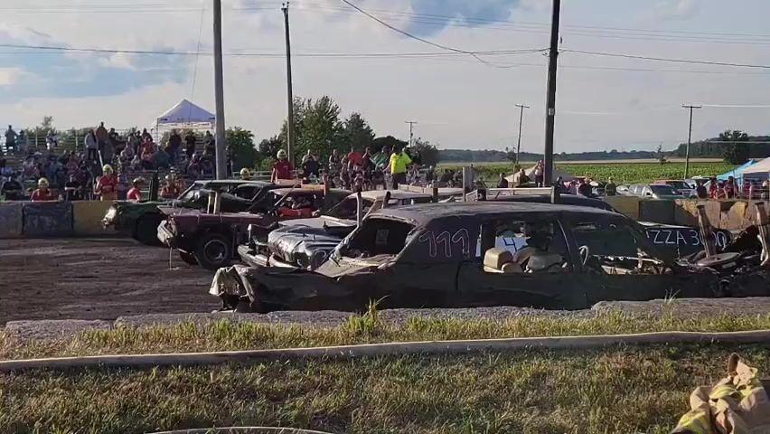 The video shows a demolition derby. Several cars, heavily modified and damaged, are lined up on a dirt track. Spectators are seated in bleachers in the background, under tents and a cloudy sky. The cars are positioned in what appears to be a starting grid, with some drivers visible inside the vehicles. The scene is set in an outdoor, rural environment.