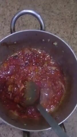 A close-up, overhead shot shows a metal pot on a countertop containing a red, glistening mixture being stirred with a metal spoon. The mixture appears to be a sauce or stew with visible onions and other ingredients. The pot has a circular rim with two metal handles on either side. The countertop has a mottled pattern.