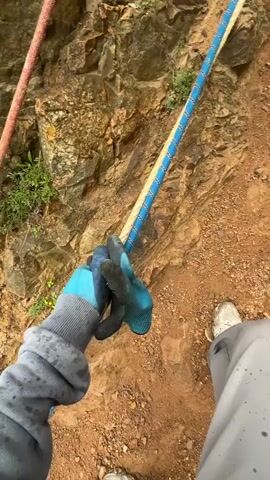 The video shows a woman climbing a steep, rocky hillside using ropes for assistance. The hillside is dry and covered in loose dirt and rocks, with some sparse vegetation. The woman is wearing a grey sweatshirt, dark pants, and light-colored shoes, and blue gloves. She holds onto the ropes to help her ascend. A person is seen climbing ahead of her. The ropes are multicolored, appearing to be red, tan and blue. In the background, the ocean is visible in the distance.