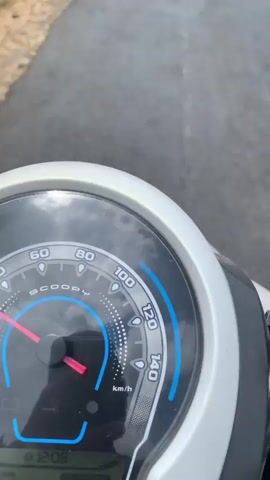 The video begins with a close-up of a motorcycle's dashboard, then pans up to show a paved road leading uphill. Green vegetation lines the sides of the road, and a wooden fence is visible on the left. In the distance, a person on a motorcycle is riding up the road, followed by a red pickup truck. The sky is blue with scattered white clouds. As the video progresses, the perspective remains on the road, and the motorcycle rider and the pickup truck continue to move further away. The scene appears to be a rural or semi-rural setting on a bright, clear day.