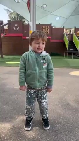 The video shows a young boy standing in a playground. He is wearing a green zip-up hooded jacket, patterned pants, and black shoes with white soles. He is holding a small stick and attempts to break it. Behind the boy is a playground structure with a pirate ship theme. It features a wooden facade, a life preserver decoration, a tic-tac-toe game, and a small set of stairs. The ground is paved with a combination of gray and green surfaces. An awning provides shade above the boy. In the background, another person can be seen on the playground structure. The boy looks at the stick and at the camera, and speaks while trying to break the stick.