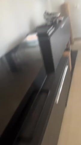 The video shows a kitten playing on a dining table. The kitten is small and has striped fur. It is initially sitting on the table, wrapped in a blanket. The kitten then stands up and moves around the table, with its tail up. The table is dark and appears to be made of wood. There are chairs around the table, and a window with blinds behind it. The room appears to be a dining room or living room.
