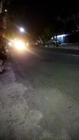 The video shows a nighttime street scene. A car's headlights illuminate the road, creating a bright glow. The road appears to be paved, with a rougher, possibly dirt or concrete, shoulder on the left. Trees and some buildings are visible in the background, dimly lit by the streetlights and the car's headlights. The overall impression is of a quiet, possibly rural or suburban street at night.