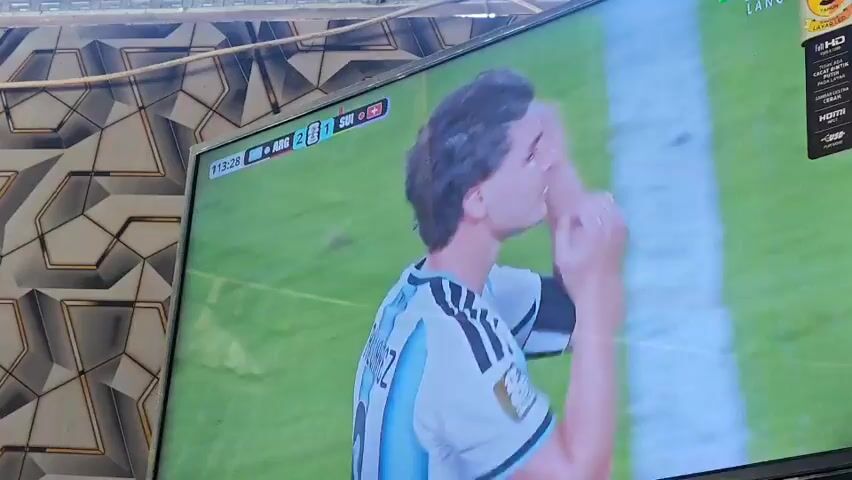 A television screen displays a soccer match. The screen shows a male soccer player wearing an Argentina jersey with the name "J. ALVAREZ" and the number "9" on the back. The player is facing away from the camera, looking towards the left side of the screen, with his left arm raised and hand extended as if signaling. A score graphic in the upper left corner shows "ARG 2" and "SUI 1", indicating the match score. A clock graphic shows "113:29". The player's jersey also has the number "1893" above his name. The background of the television screen is a green soccer field. The television is mounted on a stand, and part of the wall behind it, which has a geometric pattern, is visible.