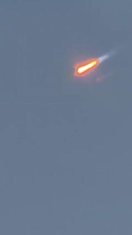 The video shows a fiery object streaking across a gray sky. The object is oval-shaped with a bright orange glow and leaves a white trail behind it. The object appears to be moving from the upper right to the lower left of the frame. The video is short, lasting only a few seconds, and the object quickly disappears from view. The sky is overcast and there are no other objects or details visible in the frame. The object's shape and trajectory suggest it could be a meteor or a falling piece of space debris.