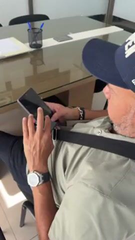 A man in a blue baseball cap, light green collared shirt, black pants, and wearing a watch and bracelet on his left wrist is seated in a chair looking at and using a black smartphone. A thick black strap goes across his chest. He is seated at a glass table with a pen holder containing pens and a piece of paper. The room is lit with bright, natural light.