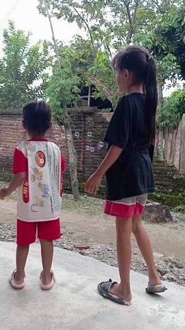 Two young girls are playing outdoors with bubbles. One girl is wearing a black shirt and pink shorts, while the other has a red and white patterned shirt and red shorts. They are both wearing sandals. They start on a concrete surface, then move onto a dirt area surrounded by trees, a brick wall, and wooden fences. The girls appear excited and are chasing and popping the bubbles. One girl holds a pink bubble wand.