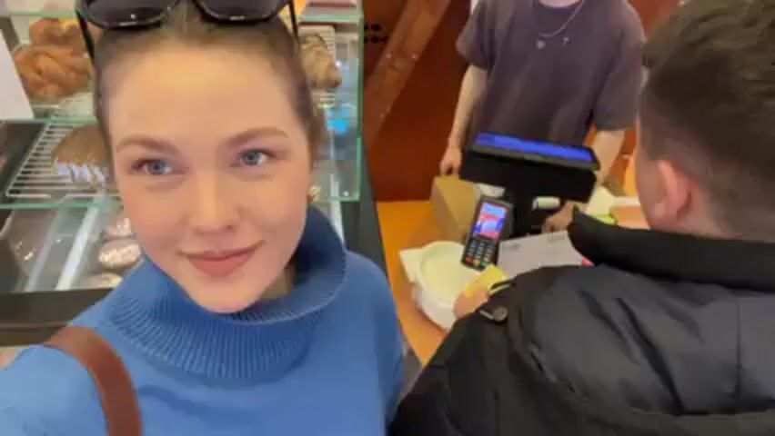 A woman wearing a blue sweater and sunglasses on her head smiles at the camera. She is standing at a bakery counter, with pastries in a glass display behind her. A man in a jacket is at the counter paying with his phone. A barista stands behind the counter. The environment is brightly lit.
