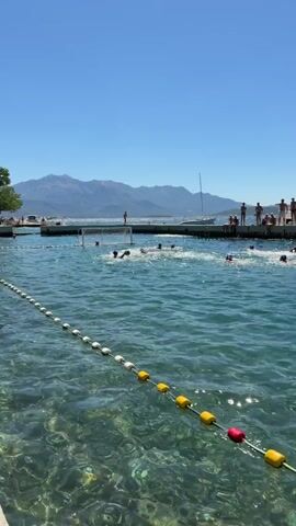 The video shows a water polo match taking place in a pool of clear blue water. The pool is outdoors, with mountains in the background under a bright blue sky. A rope with buoys separates the viewer's area from the pool. In the water, players are actively engaged in the game, with some swimming and others in the process of a play. Spectators are visible on a pier or walkway overlooking the pool. One group of spectators is seated under a covered area labeled "RESTO" and appears to be cheering or reacting to the game, with some holding up fans. Another person is seated alone on the edge of the walkway. The overall scene depicts a lively outdoor sporting event on a sunny day.