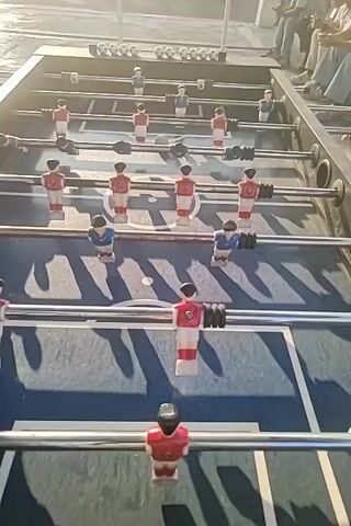 The video shows a close-up, eye-level view of a foosball table. The perspective is from behind one of the goalies, looking down the length of the table. The game is in progress, with players in red and blue jerseys positioned on the metal rods. The ball is visible in motion near the center of the table. The lighting suggests it is daytime, with shadows cast from the table and players onto the playing surface. In the background, out of focus, are people watching the game and an outdoor setting, possibly a park or recreational area, with a light-colored surface and a vertical pole visible.